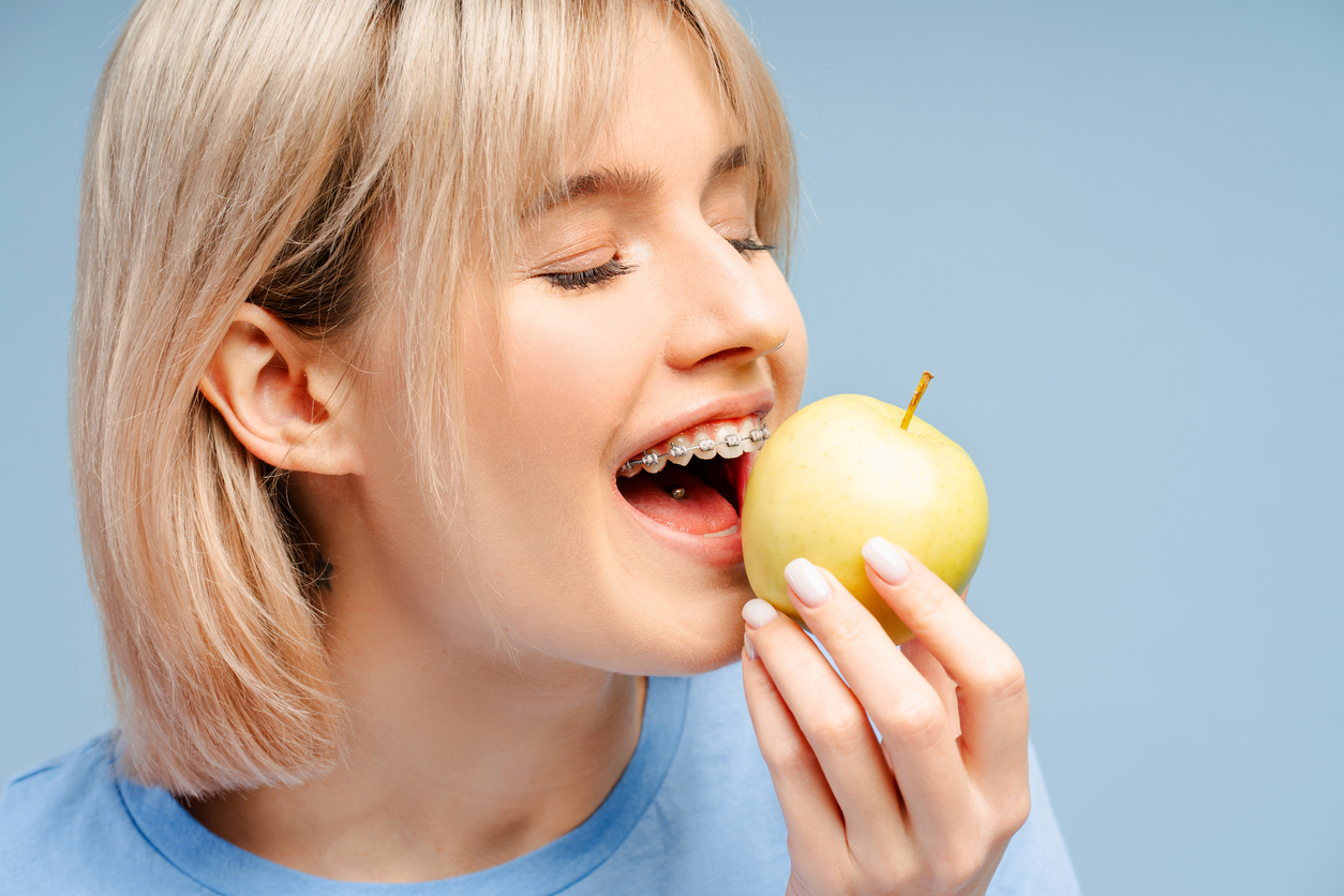 Teen with metal braces biting into a green apple against a light blue background, illustrating foods that may be hard or risky to eat with orthodontic braces.