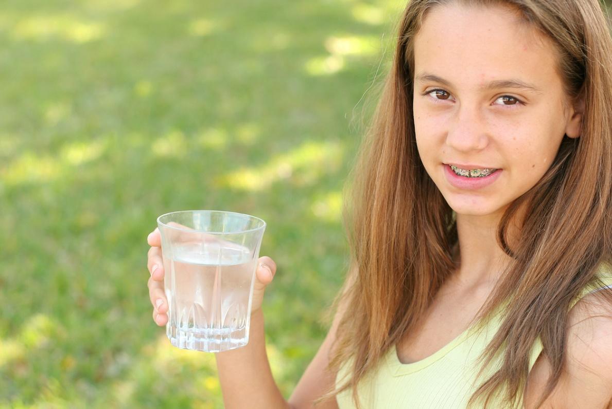 Teen with braces holding a glass of water outdoors, promoting healthy drink choices during her orthodontic treatment